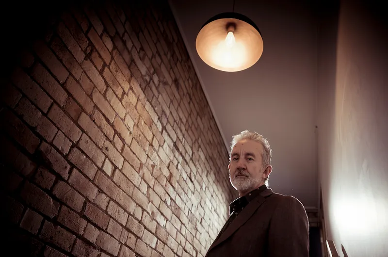 Nigel Latta next to a brick wall with gloomy lighting.