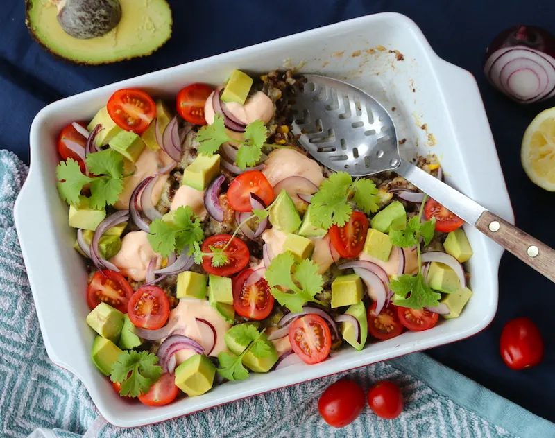 A casserole dish filled with mince, beans, and quinoa. It is topped with avocado chunks, halved cherry tomatoes, red onion slices and coriander leaves.
