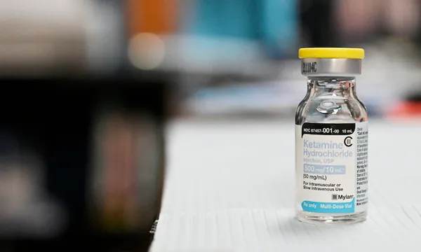 A close-up of a glass vial with a label reading ketamine. It is sitting on a white surface. The background is blurred
