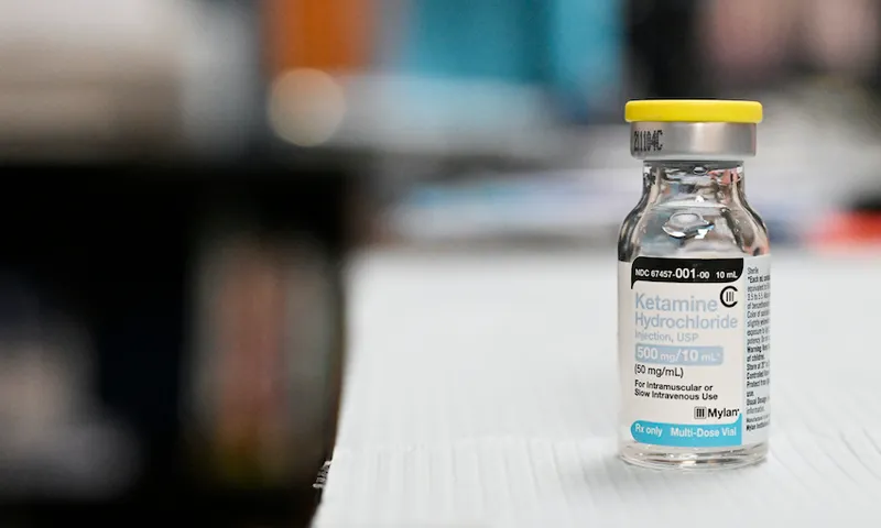 A close-up of a glass vial with a label reading ketamine. It is sitting on a white surface. The background is blurred