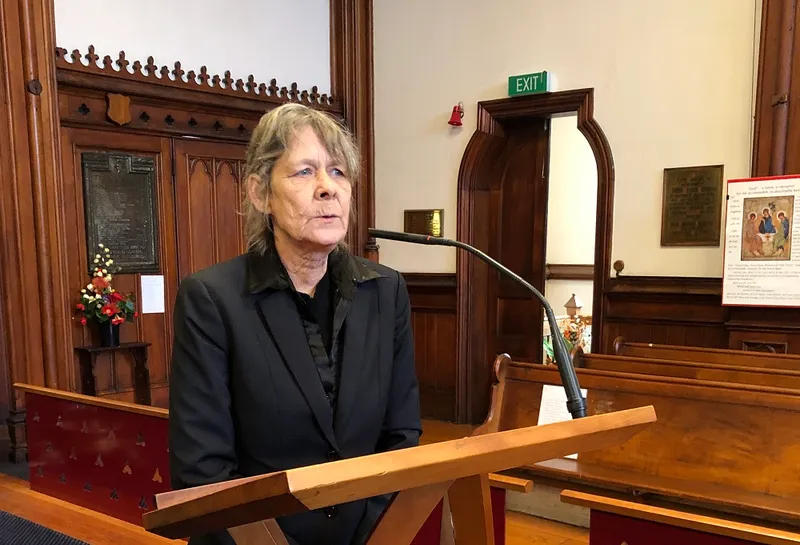an older pakeha woman wearing a sharply tailored black suit with wood panels behind her