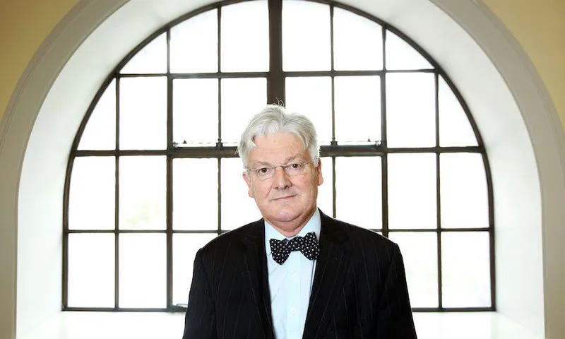 An older man with gray hair, glasses, and a bow tie stands indoors in front of a large arched window with grid-style panes, wearing a dark pinstripe suit.