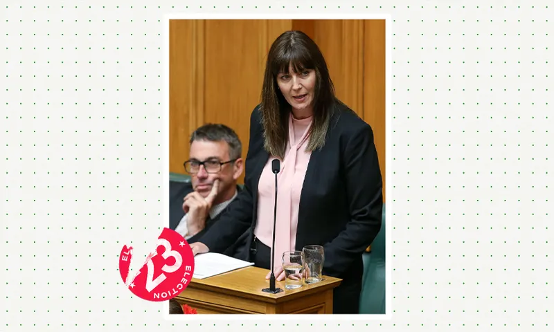 a middle aged dark heaired woman in a black blazer with someone sitting next to her against the characteristic green seats of parliament