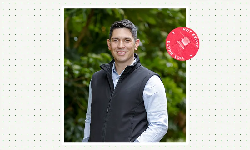 a youngish man on a green outdoors background wearing a black vest and blue shirt and smiling