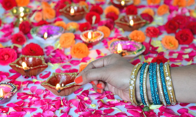 A woman's arm wearing many sparkly bangles, with painted orange nails, holds a match to light colourful diyas (small oil lamps) on a surface strewn with flower petals