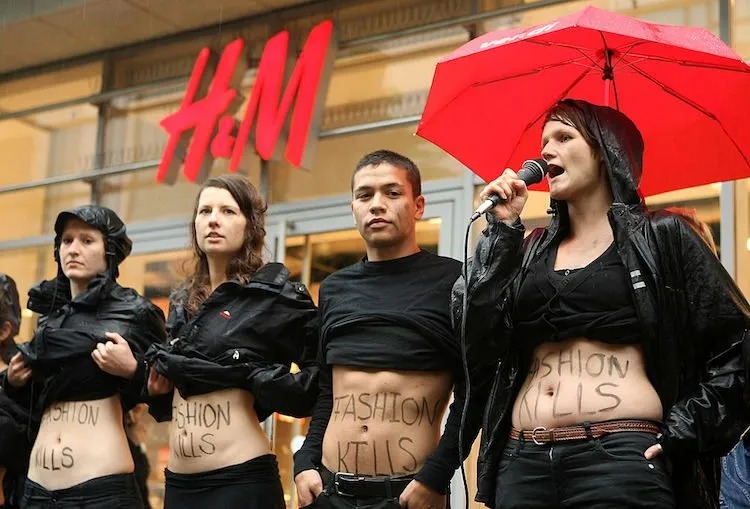 young white people wearing black standing in front of an H& M store with their black shirts hitched up to show FASHION KILLS written across their tummies