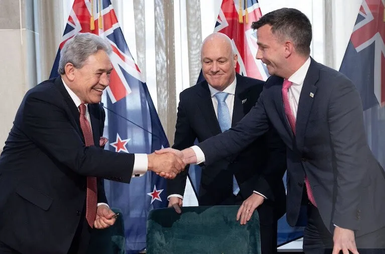 Three men in suits, standing and smiling, shake hands in front of multiple New Zealand flags. Two are shaking hands while the third man stands between them, looking on.