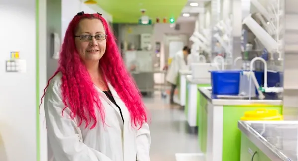 a pink haired white woman in a lab cote with a green painted lab setting in the background