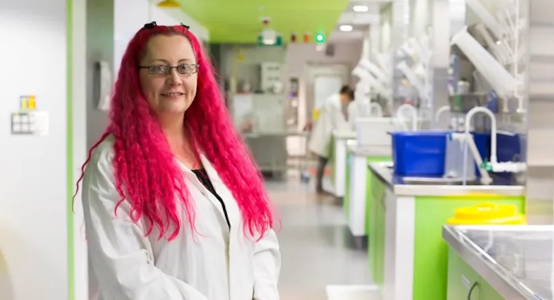a pink haired white woman in a lab cote with a green painted lab setting in the background