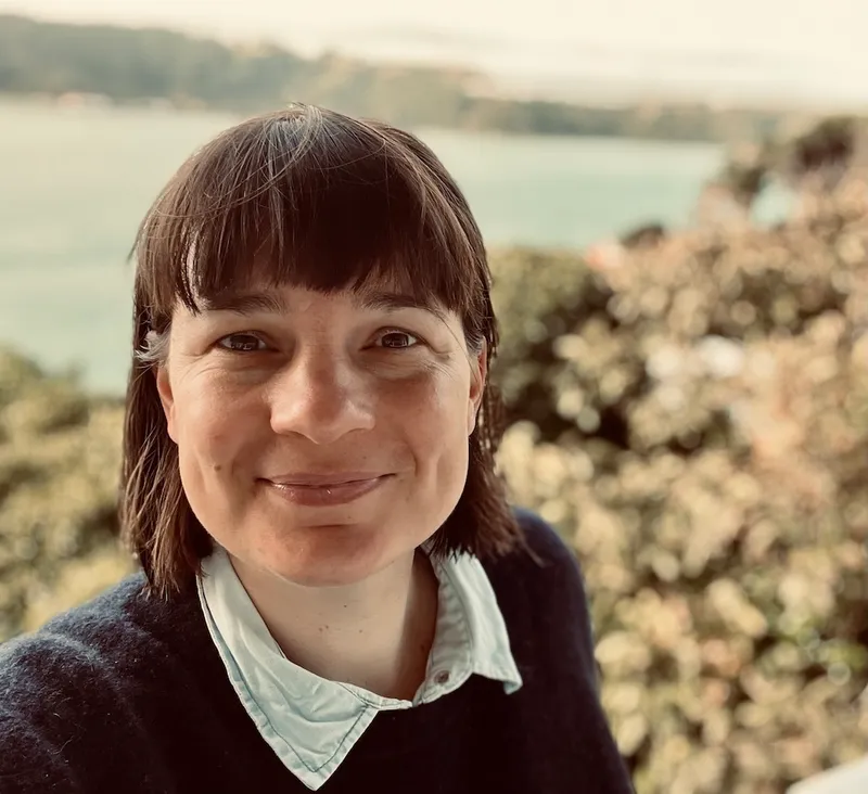 A white woman with short darck heair and collared shirt smiling at the camara with water in the background
