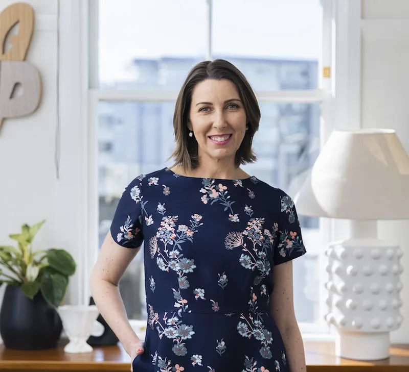Middle-aged white woman photographed in an airy apartment, houseplant, cool lamp etc.