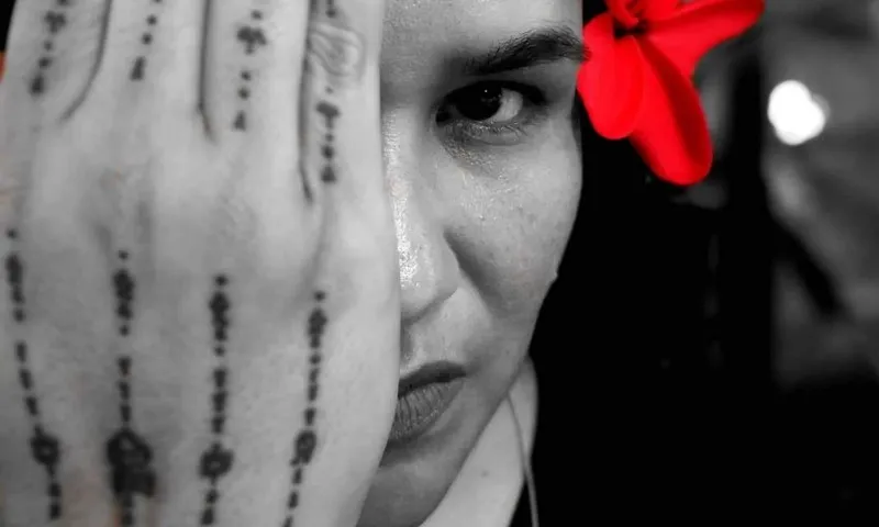 Photograph of a Samoan woman, hibiscus behind her ear, one hand over most of her face. Beautiful tatou on her hand.