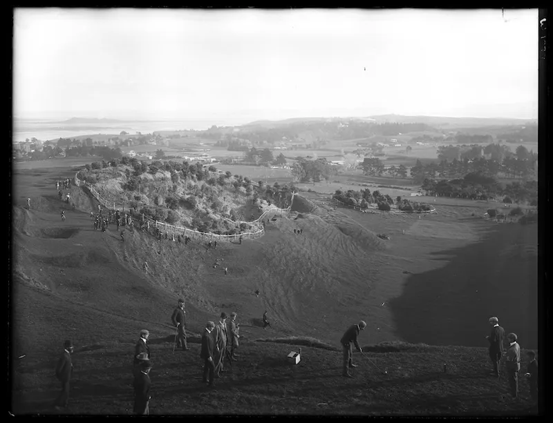 Black and white photo taken from elevated point, looking down at a crater / mountain slopes. A bunch of men are playing golf, bystanders in attendance.