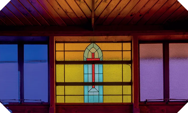 Gorgeous photograph of a stained-glass window, all yellow, purple and blue, with a crucifix picked out in the centre. Wooden ceilings soaring above.