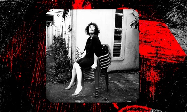 A black and white photo of a young woman sitting on a pile of plastic garden chairs, head back laughing, looking at you.