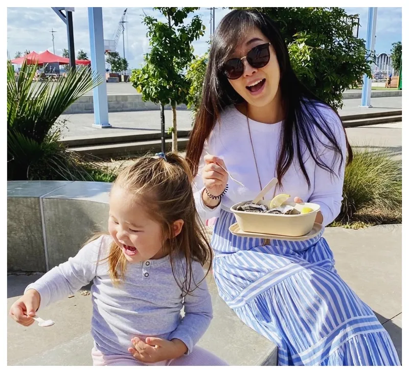 Photo of a mother and daughter perched on a wall, beautiful sunny day, tucking into kai.