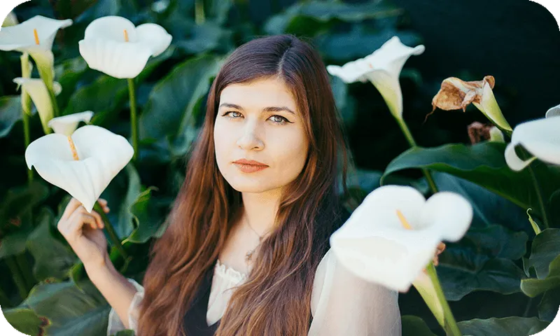 Young woman with long brown hair staring serenely at camera, backdrop of arum lilies.