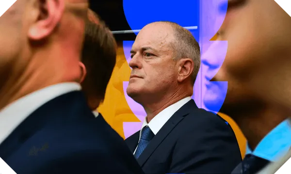 Photograph of middle-aged man in suit, staring into distance with chin lifted high. Our focus is on him but there are other suited dudes in foreground.