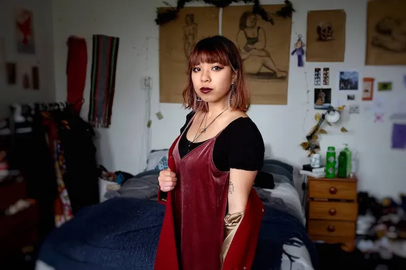Photo of young woman in her bedroom, standing front and centre, looking straight down the barrel. She wears bronze eye makeup and a velvet dress.