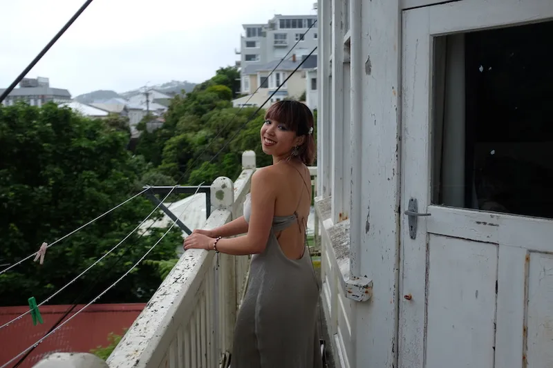 Photo of a young woman on a balcony, looks like a manky flat due to peeling paint. View of trees and buildings in background.