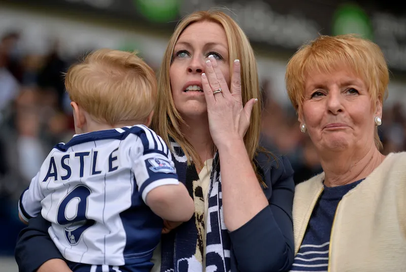 A middle-aged woman weeps as she watches a screen (out of shot, in distance). She holds a young child wearing a football jersey that reads "Astle 9"At her side is an older woman, also looking devastated.