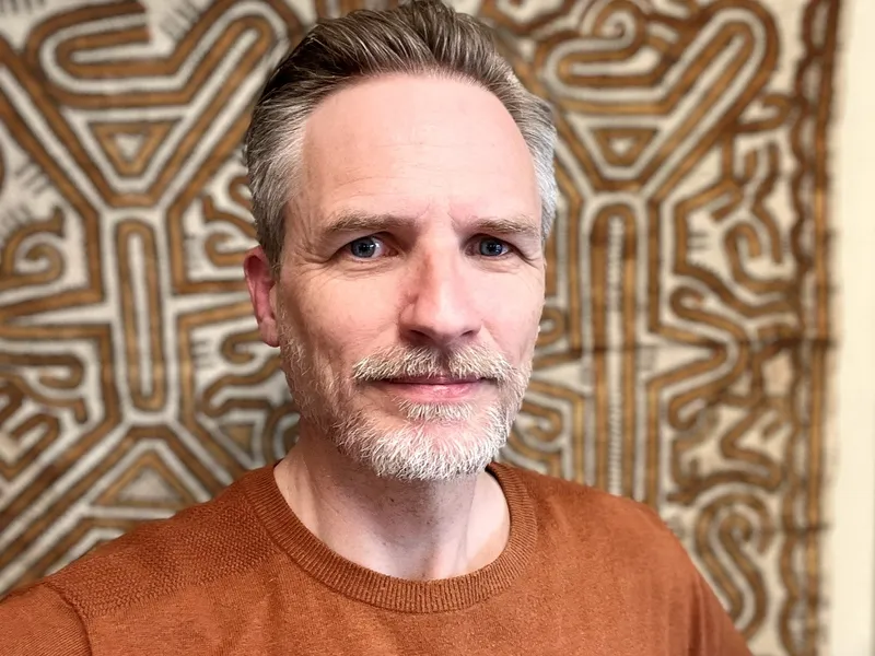 Tim thomas, a middle aged white man with a greying goatee, looks towards the camera with a small smile and a background of tapa cloth