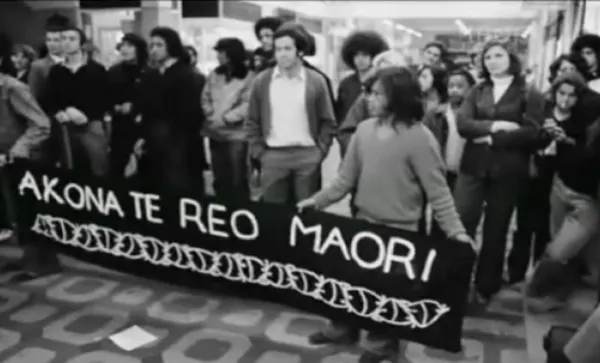 A group of people stand indoors holding a black banner reading “Akona Te Reo Maori” with a decorative border. Others stand around them, watching. The image is in black and white.