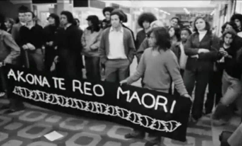 A group of people stand indoors holding a black banner reading “Akona Te Reo Maori” with a decorative border. Others stand around them, watching. The image is in black and white.