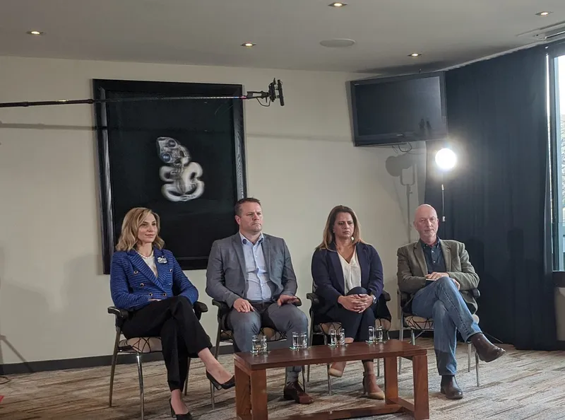 four people sitting in a row, white woman, white man in suit, brown woman in heels and blazer, bald white man in jeans.