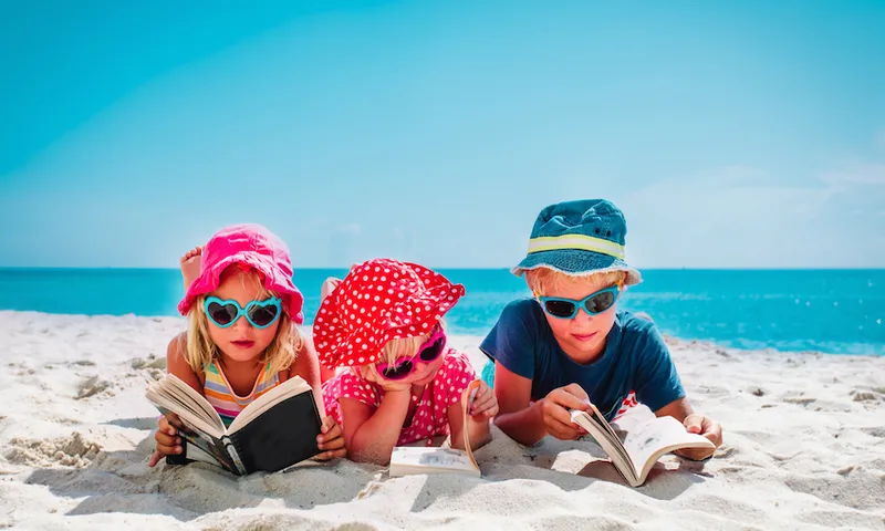Three kids lying on tummies on beach, reading books