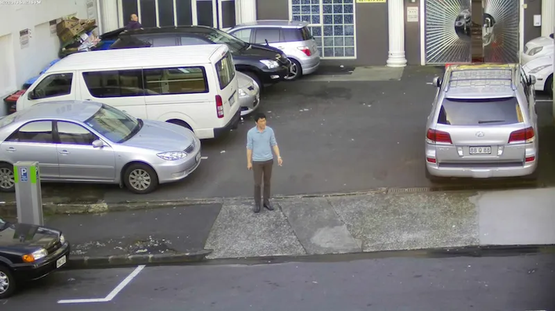 A lone man stands in a carpark, smoking, surrounded by cars.