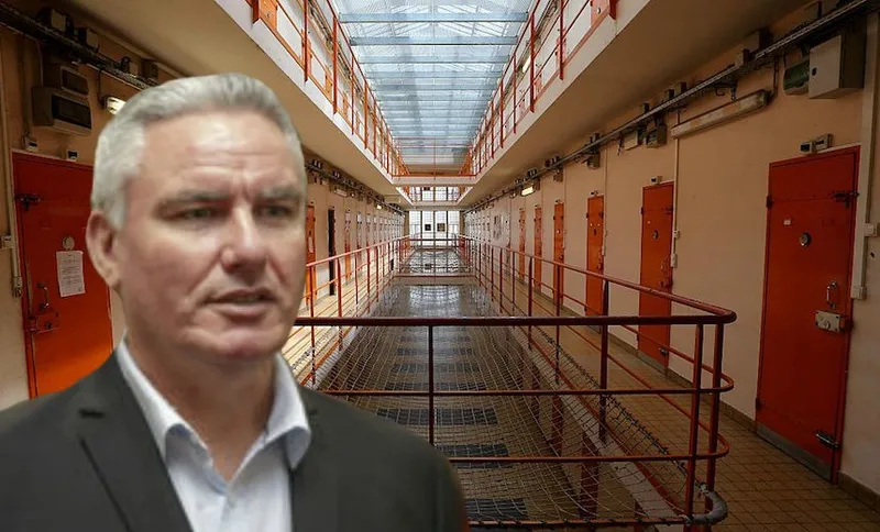 A man in a suit is in the foreground, with the background showing the interior of a prison block lined with orange cell doors and metal railings under a glass roof.