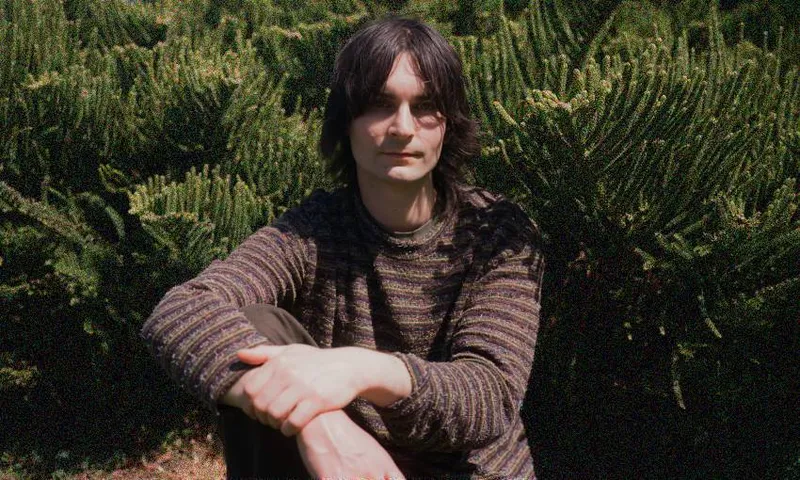 kane strang sitting in front of green bushes, hands clasped casually