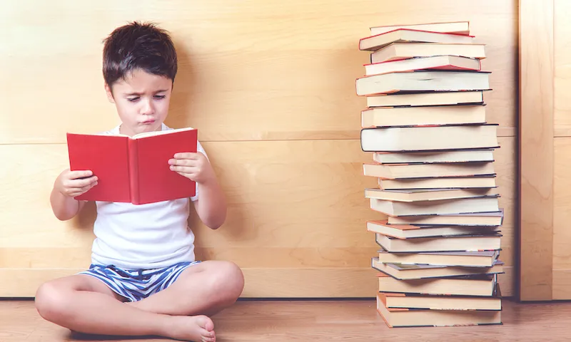 Cross-legged boy reads a book with a stack of books beside him.