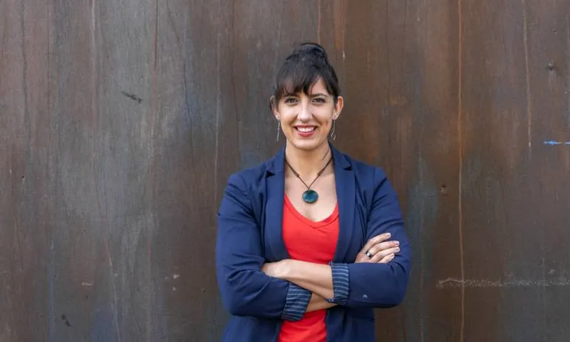 imche fourie wearing a blue blazer and red shirt, leaning against a wall with her arms crossed, smiling