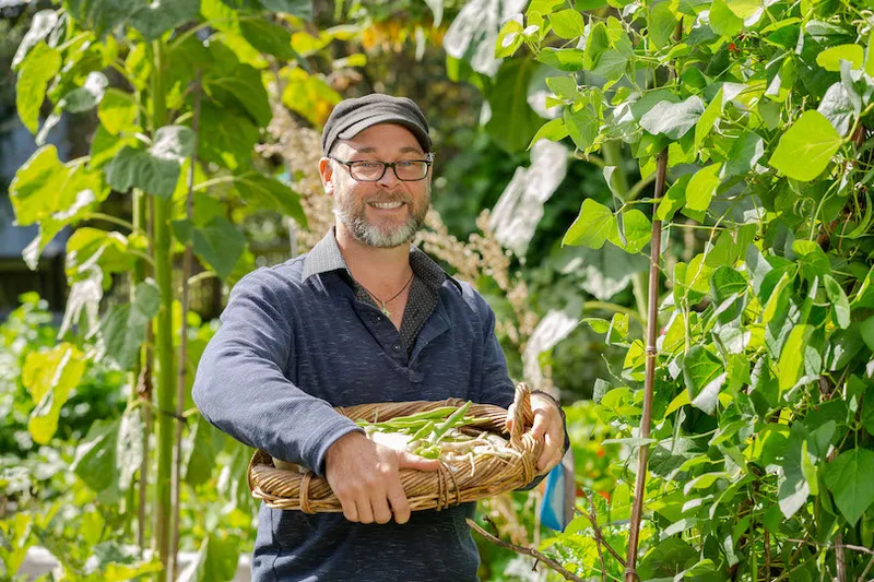 A man stands in the middle of a lush garden, holding a basket of produce, beaming.
