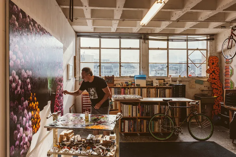 We're in an artist's studio, looking towards windows. The artist is leaning in to a huge canvas on the left-hand wall. Books and painting supplies everywhere.