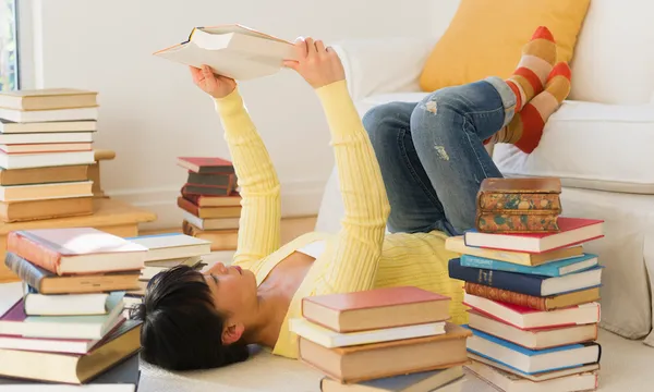 Woman lying on carpet, reading, surrounded by stacks of books.