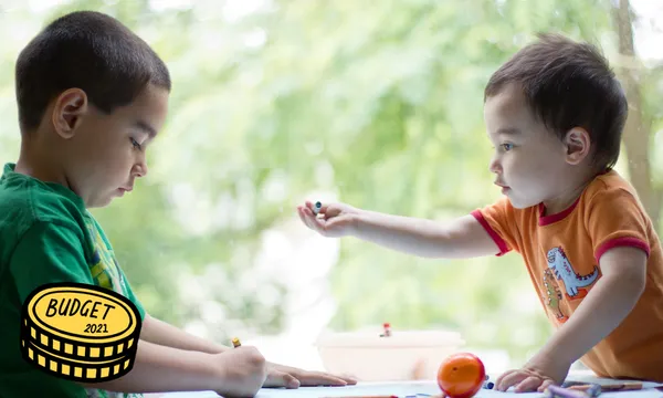 Two young mixed race boys coloring together at table next to window one year old is offering crayon to his older brother.