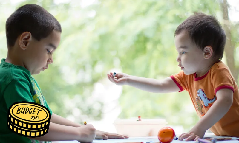 Two young mixed race boys coloring together at table next to window one year old is offering crayon to his older brother.