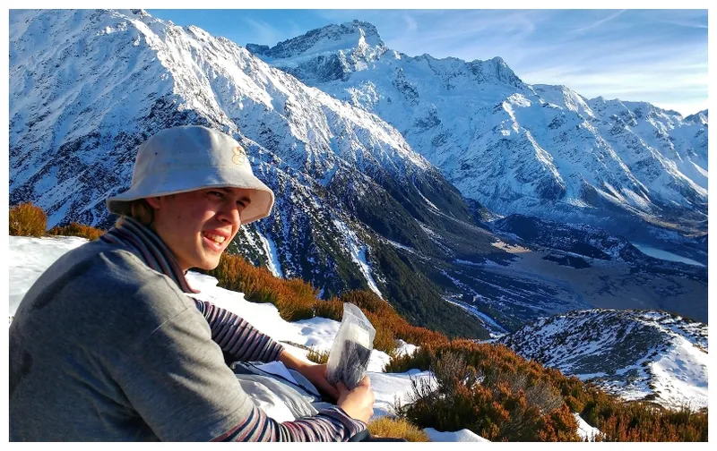 A young man sits on a snowy ridge, looking out over bare slopes and snow.