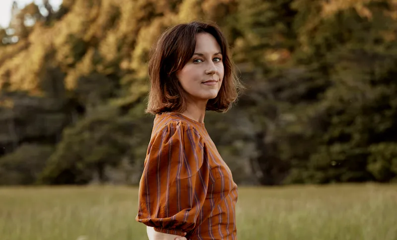 Golden hour; a middle-aged woman walks in a field, background of bush lit with sun.