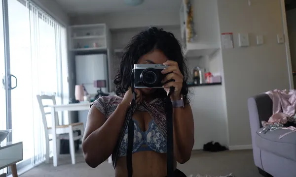 A woman stands in an apartment, wearing only a bra, face obscured by a camera.