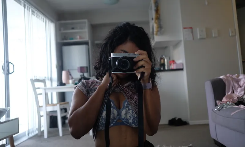 A woman stands in an apartment, wearing only a bra, face obscured by a camera.