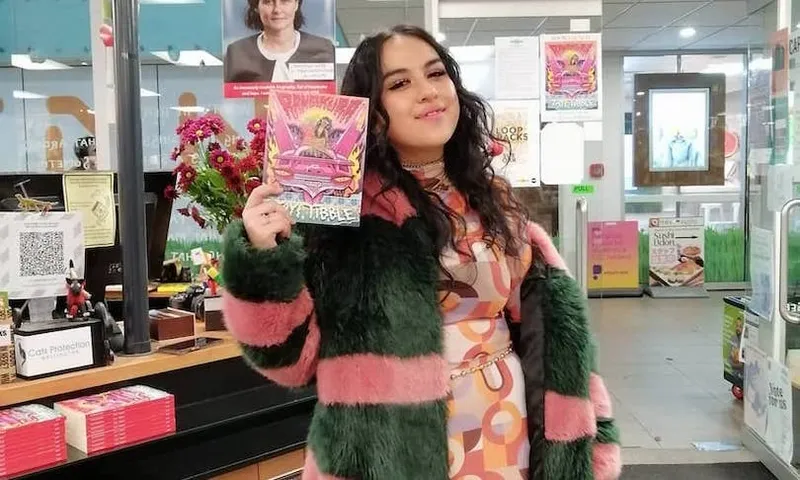 Woman in green and pink fur coat brandishes beautiful book of poetry, smiling. She's in a bookstore.