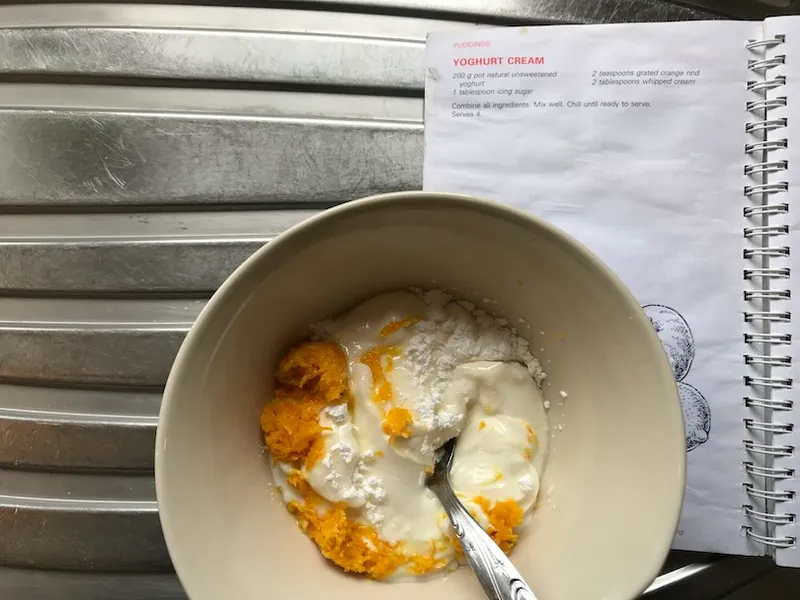 A bowl of plain yoghurt with icing sugar and orange rind on top; an Edmonds book open to Yoghurt Cream