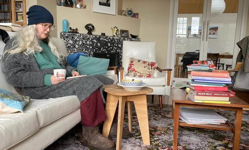 Writer Elizabeth Knox on a couch, wrapped up in a beanie and slippers, reading through a stack of books