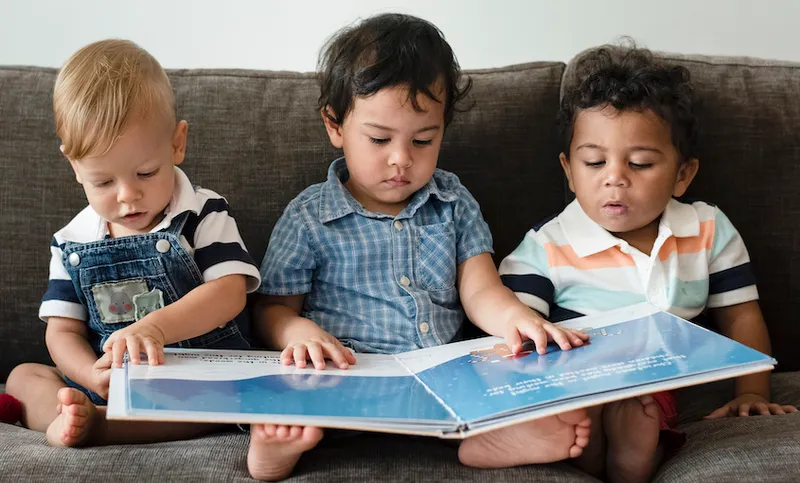 Three toddlers on a couch, reading one big picture book