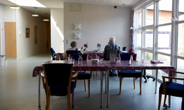 Interior of a rest home dining room; vinyl tablecloths, hard chairs, two residents seated.