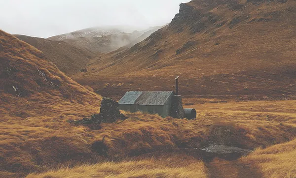 A landscape of rusty tussock, cold misty sky, a corrugated-iron hut at the base of a hill.